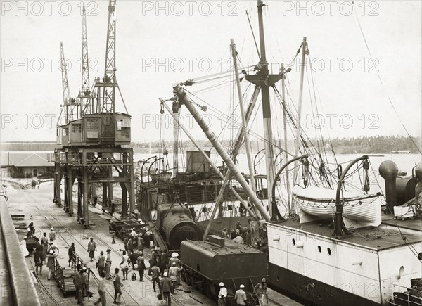 Unloading a train from SS Harmonides. Overhead view of the cranes onboard SS Harmonides lowering a locomotive boiler onto the dockside railway at Kilindini harbour, where a tender (water tank railcar or wagon) already stands. Mombasa, Kenya, circa 1926. Mombasa, Coast, Kenya, Eastern Africa, Africa.