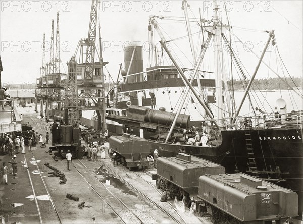 Unloading a locomotive boiler from SS Harmonides. Cranes onboard the SS Harmonides lower a train engine onto the dockside at Kilindini harbour where three tenders (water tank railcars or wagons) already stand on the dockside railway. A railway steam crane can also be seen. Mombasa, Kenya, circa 1926. Mombasa, Coast, Kenya, Eastern Africa, Africa.