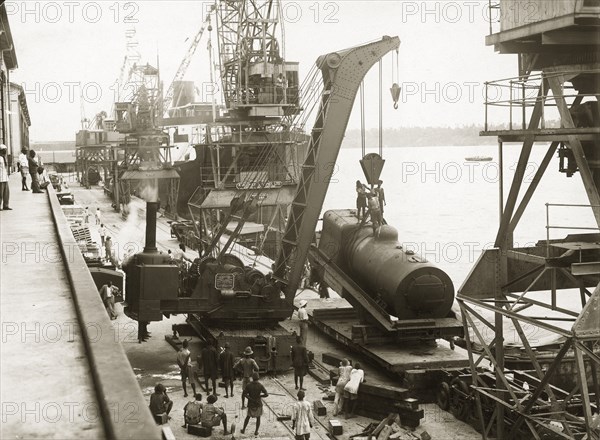 Unloading a locomotive boiler at Kilindini harbour. A railway steam crane lowers a train engine onto a bogie on the dockside at Kilindini harbour. The SS Harmonides which transported the train to Kenya is just visible, docked further along the harbour. Mombasa, Kenya, circa 1926. Mombasa, Coast, Kenya, Eastern Africa, Africa.