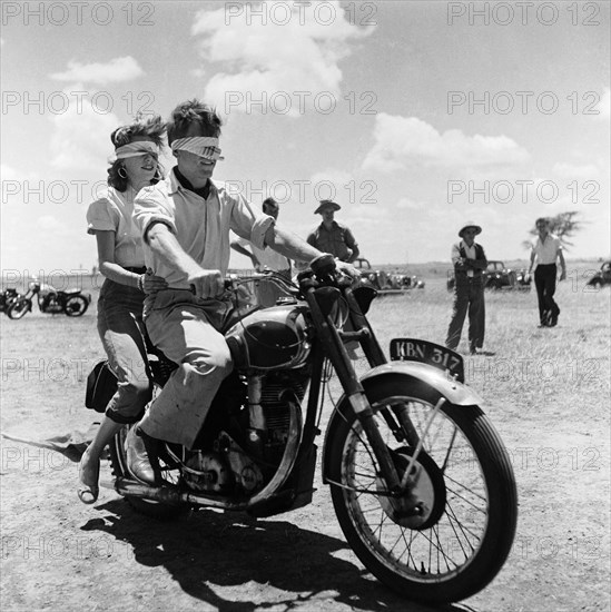 Blindfolded biking. A blindfolded couple sit astride a motorcycle at a motor sports gymkhana. Pat Dale sits in the driving seat with a female passenger riding pillion behind him. Kenya, 21 February 1954. Kenya, Eastern Africa, Africa.