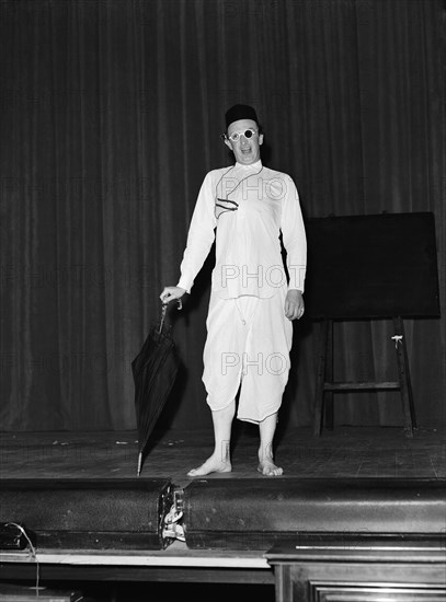 Five minute Alibhai'. Wearing theatrical costume, Peter Colmore performs his 'Five minute Alibhai' act on stage against a curtained backdrop. His props include a pair of glasses with one black lens and an umbrella. One of several acts performed in a show called 'Re-face it'. Kenya, 24 February 1953. Kenya, Eastern Africa, Africa.