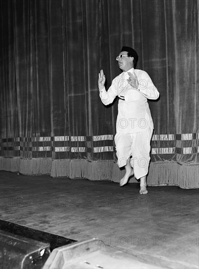 Five minute Alibhai'. Wearing theatrical costume, Peter Colmore performs his 'Five minute Alibhai' act on stage against a curtained backdrop. One of several acts performed in a show called 'Re-face it'. Kenya, 24 February 1953. Kenya, Eastern Africa, Africa.