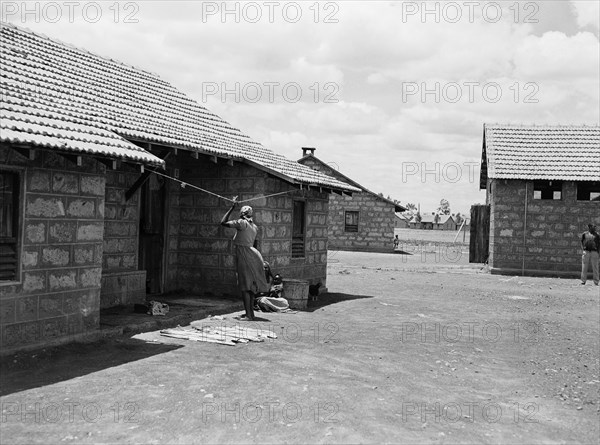 African housing. Two woman chat outside the doorway to one of three houses with pitched, tiled roofs. An original caption suggests the dwellings may have been built to house employees of Sisal Products Ltd. Kenya, 25 February 1953. Kenya, Eastern Africa, Africa.