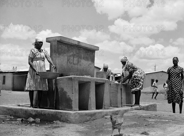 Wash place. Women wearing headscarves and patterned dresses wash bowls and collect water from taps at a communal washing place and fountain. An original caption suggests the fountain may have been built to provide water for local employees of Sisal Products Ltd. Kenya, 25 February 1953. Kenya, Eastern Africa, Africa.