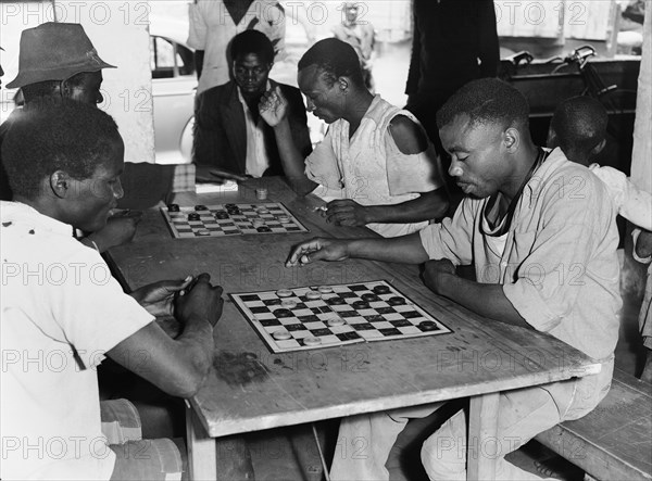 Draughts players. A group of men sit on benches around a table to play draughts. An original caption suggests they may be employees of Sisal Products Ltd. Kenya, 25 February 1953. Kenya, Eastern Africa, Africa.