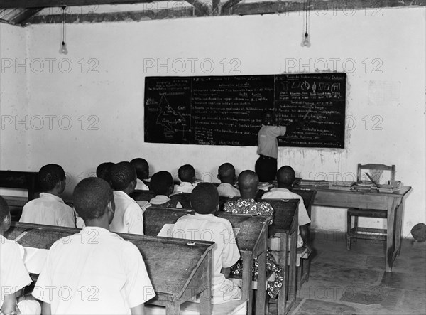 Kenyan school room. A classroom of children sit in rows behind desks listening to a teacher who is writing on a blackboard as he talks to the class. Kenya, 25 February 1953. Kenya, Eastern Africa, Africa.