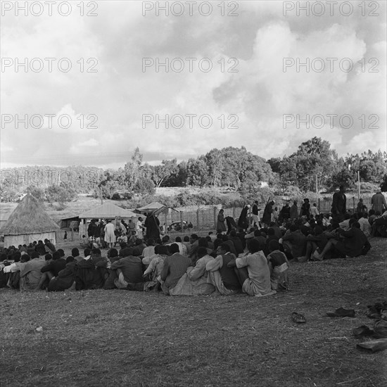 Prisoners in camp. Prisoners sit together in tightly packed lines in a fenced camp watched over by guards with guns. Uplands, Kenya, 29 March 1953. Kenya, Eastern Africa, Africa.