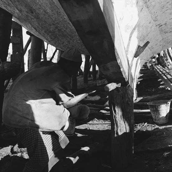 Dhow under repair. A workman hammers the underside of a dry-docked dhow. Zanzibar (Tanzania), 3-12 March 1953., Zanzibar Central/South, Tanzania, Eastern Africa, Africa.