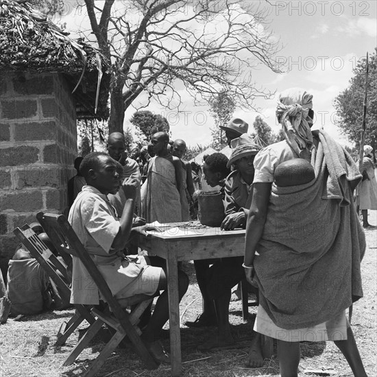 Interview board. A queue of women at a cleansing ceremony wait near a desk manned by two men equipped with pens and paper. On the other side of the desk, two men sit with coins in front of them. Kenya, 20-28 March 1953.


The two men opposite them have a bag and a collection of coins infront of them. Kenya, Eastern Africa, Africa.