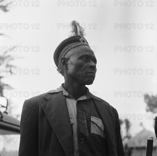 Witch doctor. Upper body shot of a witch doctor wearing a jacket and a feathered hat. Kenya, 20-28 March 1953. Kenya, Eastern Africa, Africa.
