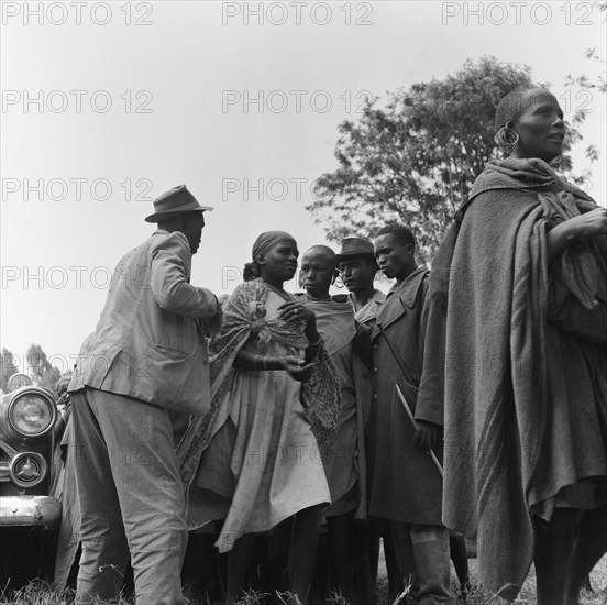 Group at a cleansing ceremony. Three men with hats and batons stand either side of a queue of women at a cleansing ceremony. Kenya, 20-28 March 1953. Kenya, Eastern Africa, Africa.