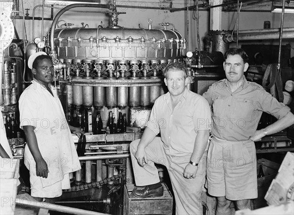 EA Breweries. Three men stand next to brewing machinery at an EA Breweries factory. The black worker pictured left holds a bottle and wears an apron printed with the letters 'EA'. Kenya, 21 April 1953. Kenya, Eastern Africa, Africa.
