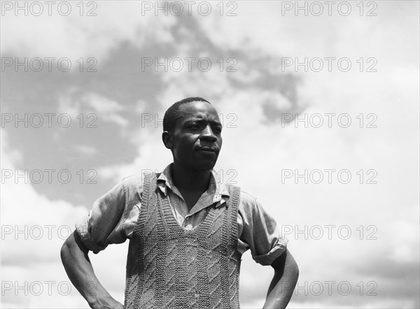Headman Samuel. Upper body shot of Headman Samuel standing, hands on hips, wearing a shirt and a knitted tank top. Kenya, 1 May 1953. Kenya, Eastern Africa, Africa.