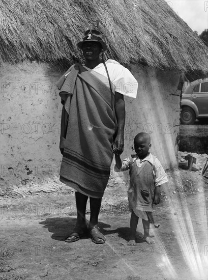 Chief Makimei. Chief Makimei holds the hand of a young boy. He wears a blanket wrapped around one shoulder and carries a shotgun on a strap over his arm. A metal badge in the shape of a lion is pinned to his hat and reads 'CHIEF'. Kenya, 12 May 1953. Kenya, Eastern Africa, Africa.