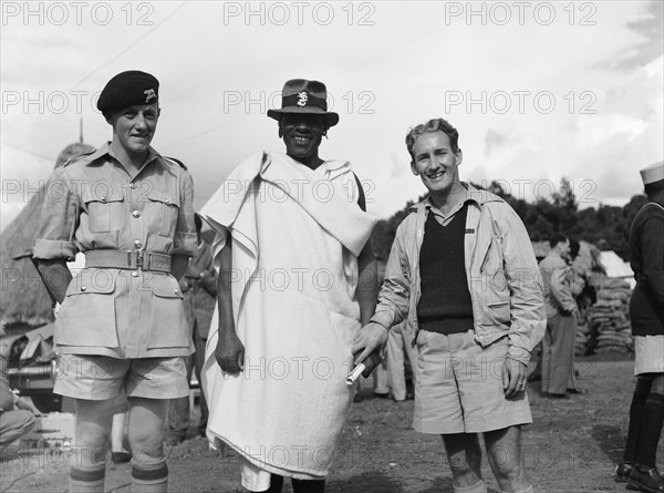 Chief Makimei with KPR officers. Chief Makimei with two Kenya Police Reserve (KPR) officers. Makimei holds a scroll of paper in one hand and wears a blanket wrapped around one shoulder. His hat displays a metal badge in the shape of a lion that reads 'CHIEF'. The KPR officer on the left wears the standard KPR uniform. Kenya, 16 May 1953. Kenya, Eastern Africa, Africa.