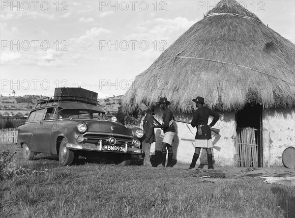 Askaris and Ford. A group of armed askaris direct a man towards a Ford car parked outside a thatched hut. Kenya, 21 May 1953. Kenya, Eastern Africa, Africa.
