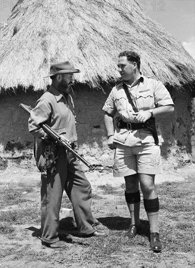 Davo' Davidson with police chief. Davo' Davidson stands outside a mud hut with a uniformed police chief. Davidson holds a large rifle under one arm and carries a revolver on his hip. Kenya, 22 May 1953. Kenya, Eastern Africa, Africa.