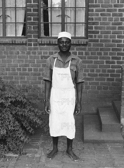 Price's room boy. The Price family's room boy pictured barefoot outside a brick building. He wears an apron that reads 'NORFOLK HOTEL' and the word 'NORFOLK' is also embroidered above his shirt pocket. Kenya, 22 May 1953. Kenya, Eastern Africa, Africa.