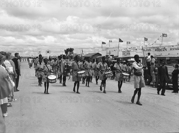 Maasai ngoma. Onlookers at a traditional 'ngoma' watch as a group of young Maasai people wearing uniforms pass through the centre of town playing drums. Kajiado, Kenya, 2 June 1953. Kajiado, Rift Valley, Kenya, Eastern Africa, Africa.