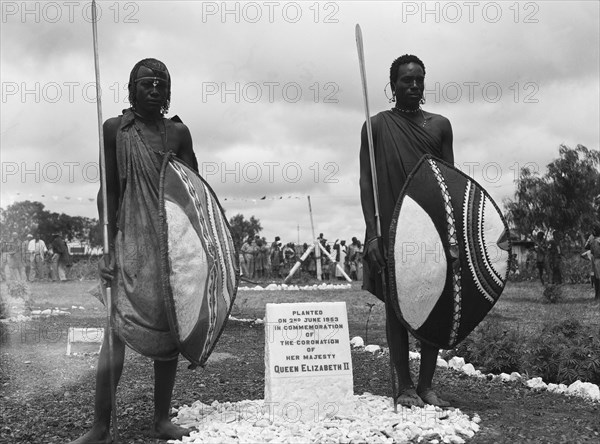 Maasai beside a coronation plaque. Maasai warriors stand either side of a memorial plaque commemorating the coronation of Queen Elizabeth II at a 'ngoma', a word meaning drum that is also used to describe traditional celebrations. They wear traditional dress and hold spears and shields. Kajiado, Kenya, 2 June 1953. Kajiado, Rift Valley, Kenya, Eastern Africa, Africa.