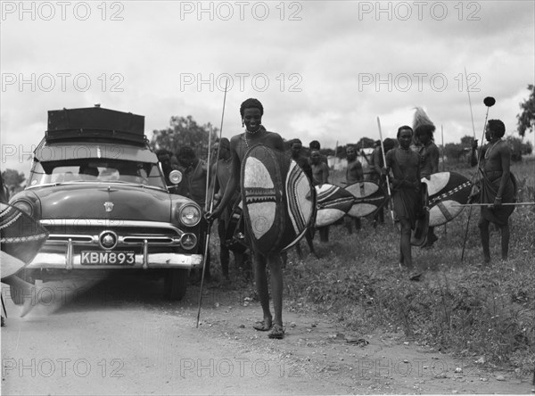 Maasai and Ford. Maasai warriors holding spears and shields congregate on the roadside next to a Ford car at a 'ngoma', a word meaning drum that is also used to describe traditional celebrations. Kajiado, Kenya, 2 June 1953. Kajiado, Rift Valley, Kenya, Eastern Africa, Africa.