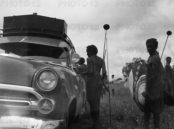 Maasai and Ford. Maasai warriors holding spears and shields congregate on the roadside near a Ford car at a 'ngoma', a word meaning drum that is also used to describe traditional celebrations. Kajiado, Kenya, 2 June 1953. Kajiado, Rift Valley, Kenya, Eastern Africa, Africa.