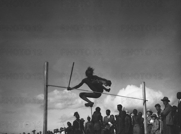 High jump. A man holding a baton attempts the high jump at a 'ngoma', a word meaning drum that is also used to describe traditional celebrations. Kajiado, Kenya, 2 June 1953. Kajiado, Rift Valley, Kenya, Eastern Africa, Africa.