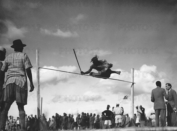 High jump. A man holding a baton attempts the high jump at a 'ngoma', a word meaning drum that is also used to describe traditional celebrations. Kajiado, Kenya, 2 June 1953. Kajiado, Rift Valley, Kenya, Eastern Africa, Africa.