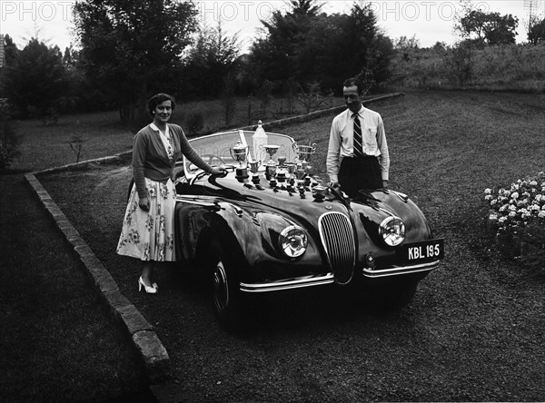 Manussis with trophies. Racing driver John Manussis poses with a woman next to a Jaguar Coupe convertible car, the bonnet of which is displays numerous trophies. Kenya, 7 June 1953. Kenya, Eastern Africa, Africa.