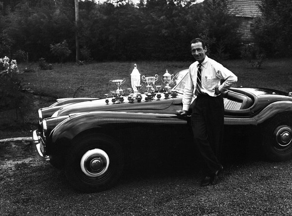 Manussis with trophies. John Manussis leans on the wing of a Jaguar Coupe convertible car, the bonnet of which displays numerous trophies. Kenya, 7 June 1953. Kenya, Eastern Africa, Africa.