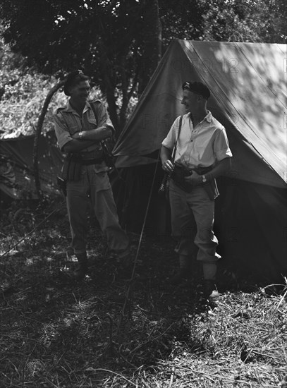 Uniformed soldiers at a KAR camp. Two armed and uniformed European soldiers share a joke at a bivouac set up by the King's African Rifles (KAR). Both men wear berets displaying the insignia of the Kenya Regiment, a voluntary territorial force established in 1937 in the name of King George VI. Kenya, June 1953. Kenya, Eastern Africa, Africa.