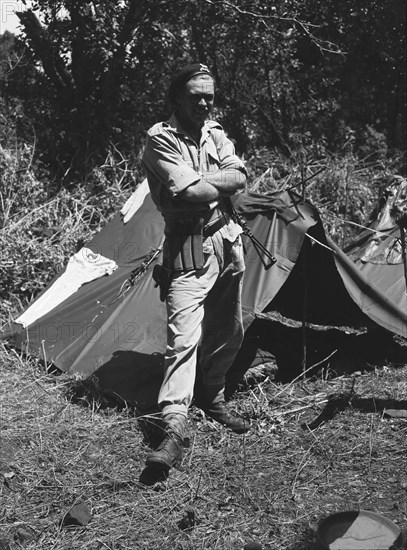 Uniformed soldier at a KAR camp. A uniformed European soldier stands outside a tent at a bivouac set up by the King's African Rifles (KAR). He wears a beret displaying the insignia of the Kenya Regiment, a voluntary territorial force established in 1937 in the name of King George VI. Kenya, June 1953. Kenya, Eastern Africa, Africa.