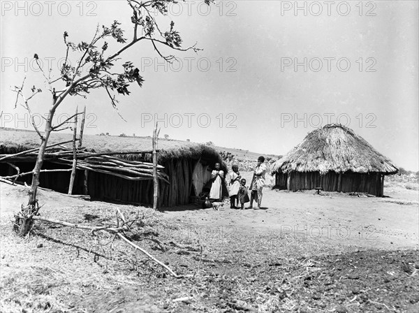 Mbulu hut. An adult and three children stand outside a long wooden hut with a flat, thatched roof. Kenya, August 1953. Mbulu, Rift Valley, Kenya, Eastern Africa, Africa.