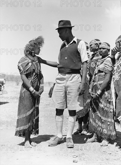 Barabaig chief with young women. A chief from the Barabaig tribe looks down on a young Barabaig woman wearing traditional dress. Other young women can be seen to the right, their clothes heavily decorated with beaded patterns and tassles: their heads, hands and feet adorned with ornate jewellery. Probably Tanganyika Territory (Tanzania), August 1953. Tanzania, Eastern Africa, Africa.