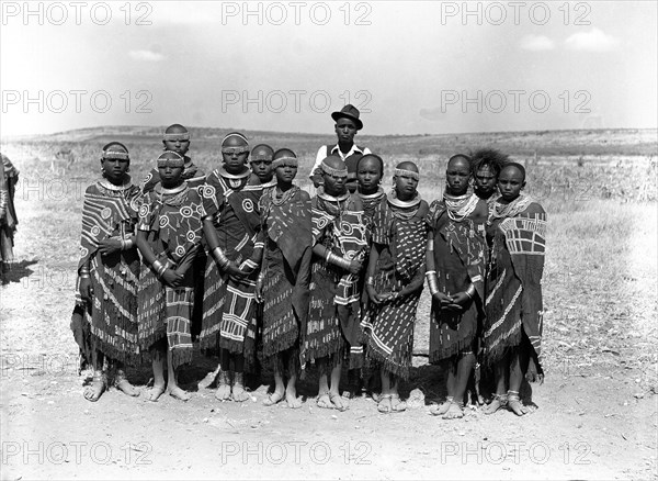Barabaig chief with young women. A chief from the Barabaig tribe stands over a group of young Barabaig women who wear traditional dress. Their clothes are heavily decorated with beaded patterns and tassles: their heads, hands and feet are adorned with ornate jewellery. Probably Tanganyika Territory (Tanzania), August 1953. Tanzania, Eastern Africa, Africa.