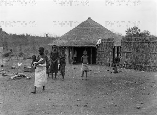 Mozambique natives. A group of children outside a wooden hut with a thatched roof. A barefooted woman in the foreground carries a baby on her hip as she walks. Mozambique, September 1953. Mozambique, Southern Africa, Africa.
