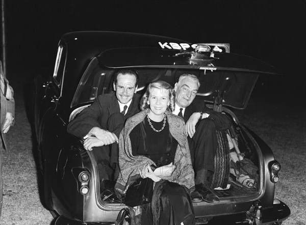 In the boot. Two men wearing suits sit hunched up in the open boot of a car behind a woman wearing an evening dress at the Brunton's party. Kenya, 14 February 1953. Kenya, Eastern Africa, Africa.