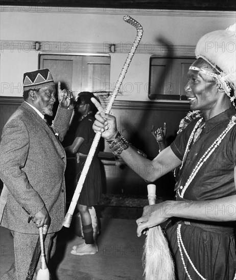Jomo Kenyatta salutes a female bodyguard. An African man wearing traditional dress in the foreground overshadows Jomo Kenyatta, the new leader of independent Kenya, as Kenyatta is captured exchanging a 'one for all' salute with a member of his female bodyguard on a railway platform. Kenya, circa 1963. Kenya, Eastern Africa, Africa.