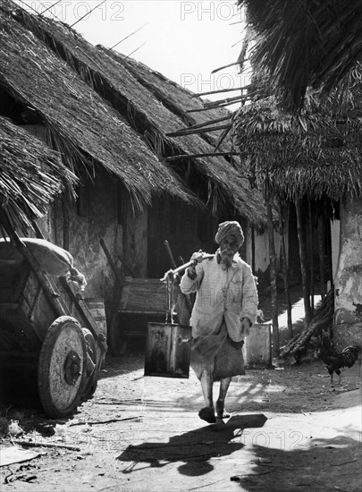 Street scene in old Mombasa. An Indian man, barefoot and wearing a turban, walks down a narrow, unpaved street with two kerosene cans slung on a pole across his shoulder. Hand carts line the street. Mombasa, Kenya, circa 1950. Mombasa, Coast, Kenya, Eastern Africa, Africa.