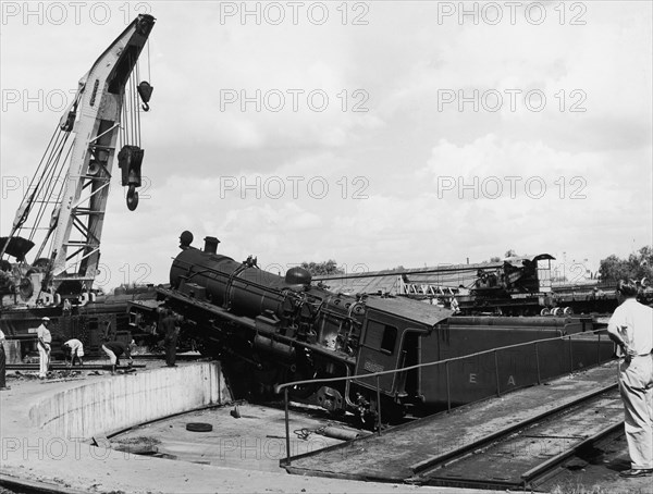 Runaway locomotive. An official photograph from the East African Railways and Harbours Administration (EAR&H) showing a locomotive boiler and tender which have apparently overshot the line and mounted a platform. A crane is being positioned to lift the train. East Africa, probably Kenya, circa 1950.