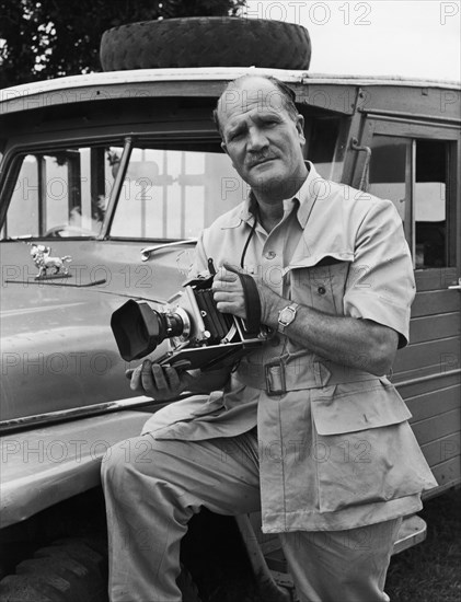 Portrait of a photographer. A publicity photograph from the East African Railways and Harbours Administration (EAH&R) features a European man, presumably an official EAH&R photographer, posing by a car holding a large camera. Kenya, circa 1950. Kenya, Eastern Africa, Africa.