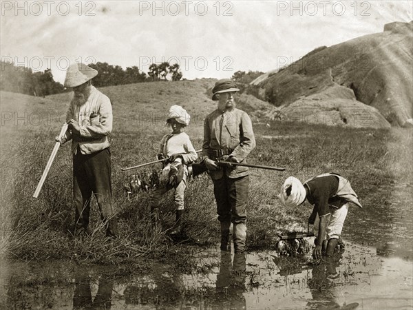 Snipe hunting. Two British snipe hunters stand, shotguns in hand, as their young Indian assistants gather downed birds and hang them onto wooden racks. India, circa 1860. India, Southern Asia, Asia.