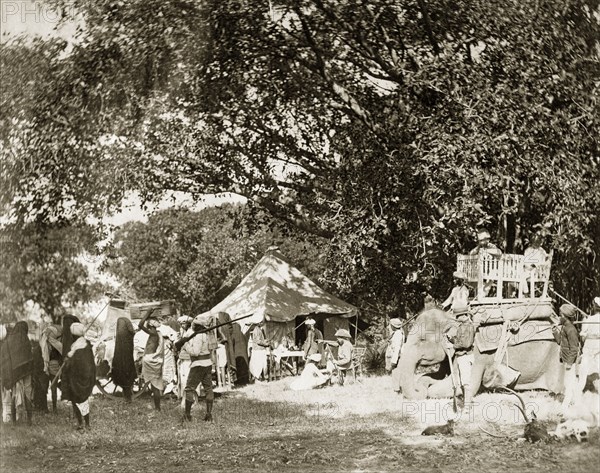 Leaving for the tiger hunt. The campsite of a hunting party bustles with activity in preparation for a tiger hunt. A number of Indian servants head off with supplies whilst two British gentlemen prepare to depart in a howdah secured to the back of an elephant. India, circa 1885. India, Southern Asia, Asia.