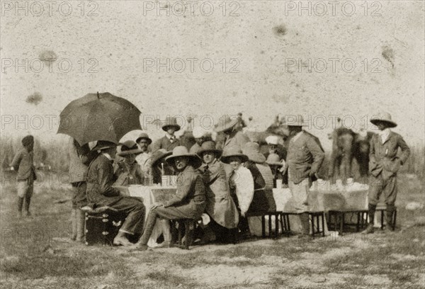 Hunting party lunch. A hunting party comprised of British men and women seek shade under solatopi hats and parasols as they eat their lunch seated around a long table. North East India, circa 1890. India, Southern Asia, Asia.
