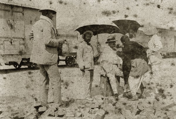 A shave in the shade. A British gentlemen is shaded with parasols as he is shaved by an Indian servant near a railway track. North East India, circa 1890. India, Southern Asia, Asia.