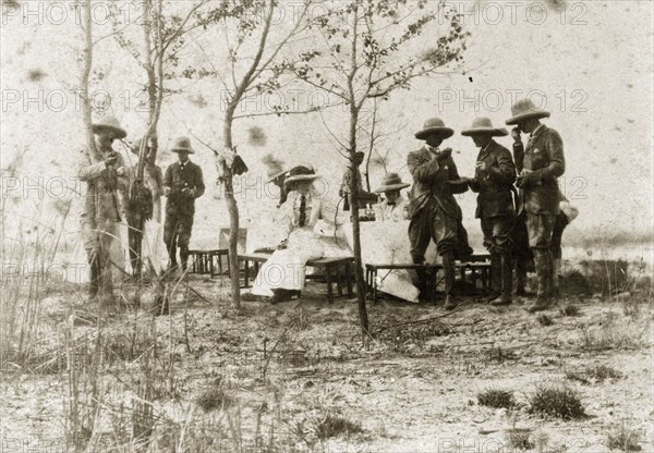 Hunting party lunch. A hunting party comprised of British men and women takes a midday break to eat lunch around a table with benches. North East India, circa 1890. India, Southern Asia, Asia.