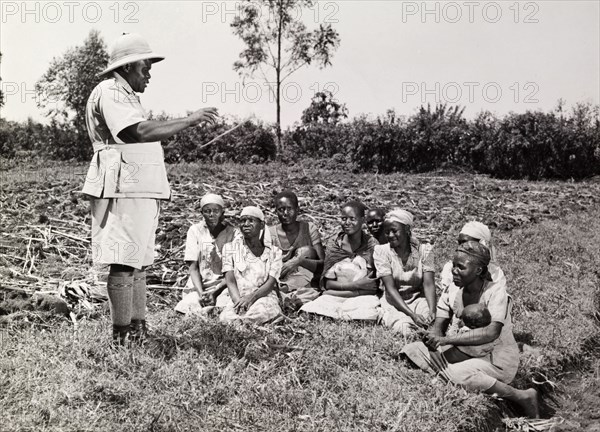 Farming advice for women. A man in uniform dispenses advice on improved farming methods to a small audience of female farmworkers. The women are seated on the ground, some holding babies. Kenya, circa 1965. Kenya, Eastern Africa, Africa.