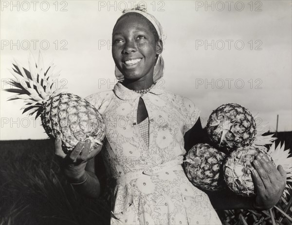 Pineapples for the canning factory. A young agricultural worker smiles for the camera as she carries an armful of pineapples that have been grown for a local canning factory. Central Province, Kenya, circa 1965., Central (Kenya), Kenya, Eastern Africa, Africa.