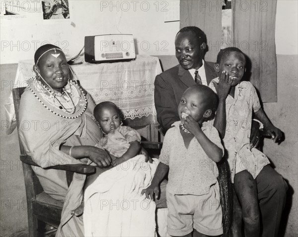 A 'modern' Maasai family. Portrait of a 'modern' Maasai family inside a home decorated with Western-style furnishings including a small radio. The mother is the only family member who retains her traditional Maasai clothing and jewellery. Kenya, circa 1965. Kenya, Eastern Africa, Africa.