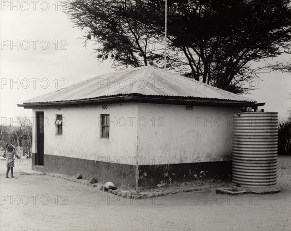 A permanent Maasai home. Portrait of a permanent Maasai home, a square building with a pitched roof and small windows. A corrugated iron container, presumbaly a water storage tank, is also visible. Kenya, circa 1965. Kenya, Eastern Africa, Africa.